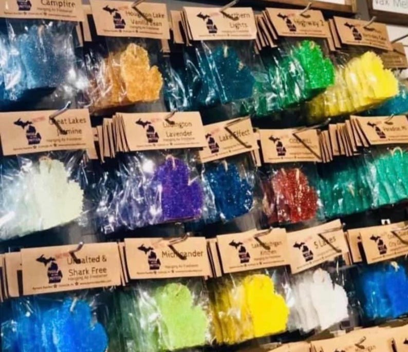 Op de afbeelding: Een display van luchtverfrissers in de vorm van de staat Michigan, in verschillende kleuren. Elk is verpakt in een doorzichtige plastic zak en bevestigd aan een bruin papieren label met tekst. De geuren zijn onder meer Campfire en Houghton Lake Vanilla Bean.