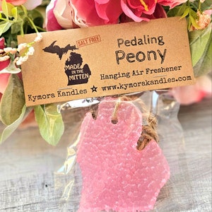 Op de afbeelding: Een roze, Michigan-vormige luchtverfrisser in een doorzichtige plastic zak, opgehangen aan een touwtje. Het label luidt "Pedaling Peony" en "Hanging Air Freshener" met de website kymorakandles.com. De achtergrond toont roze en witte bloemen.