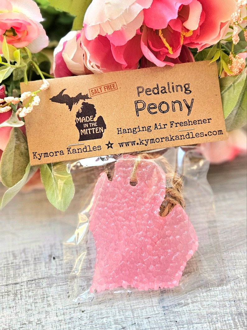 Op de afbeelding: Een roze, Michigan-vormige luchtverfrisser in een doorzichtige plastic zak, opgehangen aan een touwtje. Het label luidt "Pedaling Peony" en "Hanging Air Freshener" met de website kymorakandles.com. De achtergrond toont roze en witte bloemen.
