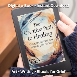 Peut inclure: Une liseuse noire affiche la couverture de "The Creative Path to Healing". La couverture présente des illustrations à l'aquarelle de plumes, d'une bougie et de formes abstraites en bleu, orange et marron. Le texte comprend "Art • Writing • Rituals for Grief."