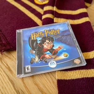 May include: A clear plastic case containing the "Harry Potter and the Sorcerer's Stone" PC game. The cover art features Harry Potter on a broomstick. The game is set on a maroon and gold striped scarf.