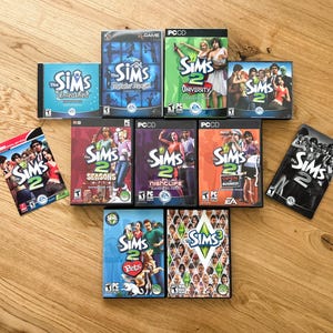 May include: A collection of The Sims PC game boxes, including The Sims 2 and The Sims 3. The boxes feature various cover art with people and game titles like "University", "Seasons", and "Pets". The games are arranged on a wooden surface.
