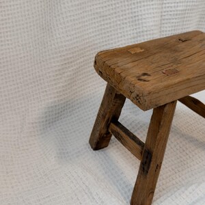 May include: A small, rustic wooden stool with a rectangular seat and angled legs. The stool is made of weathered wood, showcasing natural grain and knots. The design is simple and functional, suitable for various uses.