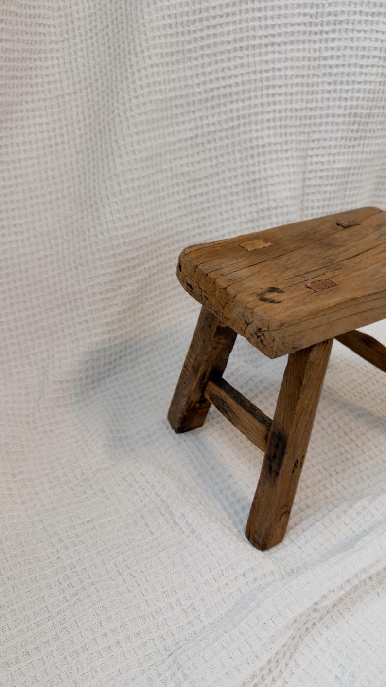 May include: A small, rustic wooden stool with a rectangular seat and angled legs. The stool is made of weathered wood, showcasing natural grain and knots. The design is simple and functional, suitable for various uses.