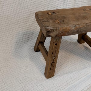 May include: A small, rustic wooden stool with a rectangular seat and three angled legs. The stool is made of weathered wood, showcasing natural grain and knots. The stool measures approximately 12 inches long, 8 inches wide, and 10 inches high.