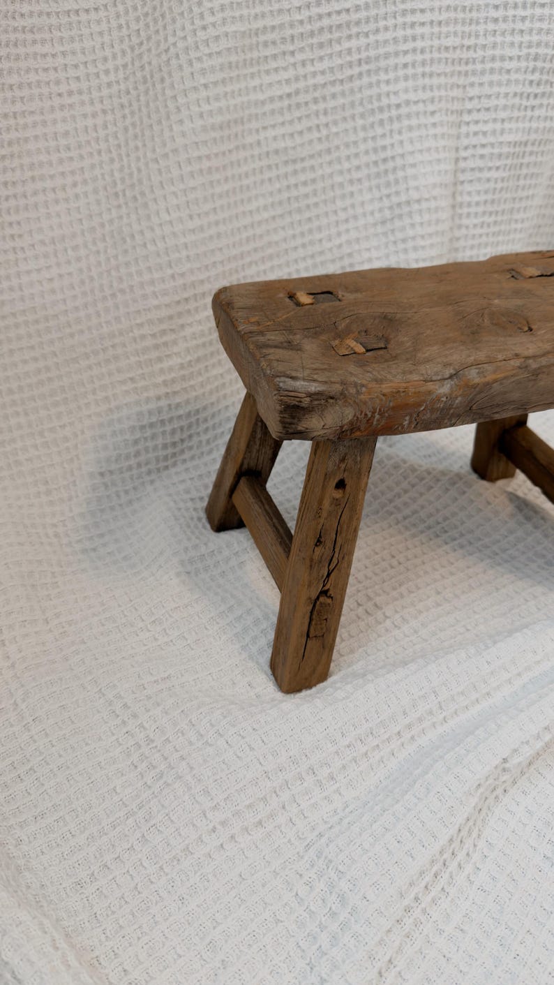 May include: A small, rustic wooden stool with a rectangular seat and three angled legs. The stool is made of weathered wood, showcasing natural grain and knots. The stool measures approximately 12 inches long, 8 inches wide, and 10 inches high.