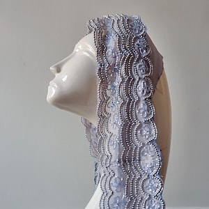May include: A light blue lace veil with silver beaded trim is draped over a white mannequin head. The veil's edge features a scalloped design. The background is a plain white wall.