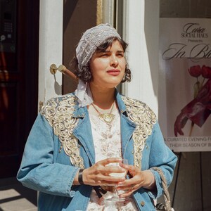 May include: A woman wearing a light blue denim jacket with gold embroidery, a white dress, and a beaded headscarf. She holds a glass of a light-colored beverage. A small, silver, geometric handbag hangs from her arm. A poster in the background reads "In Bloom."