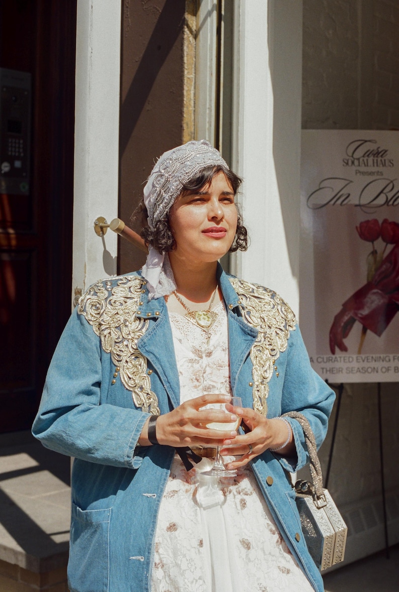 May include: A woman wearing a light blue denim jacket with gold embroidery, a white dress, and a beaded headscarf. She holds a glass of a light-colored beverage. A small, silver, geometric handbag hangs from her arm. A poster in the background reads "In Bloom."