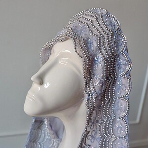 May include: A white ceramic bust wearing a light blue veil with silver beaded trim. The veil is intricately designed with floral patterns and a scalloped edge. The bust is smooth and glossy, with a serene expression.