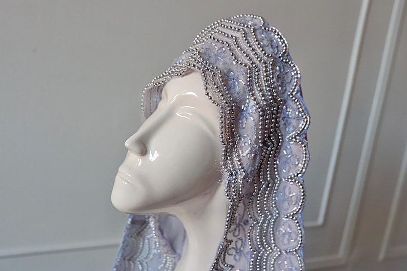 May include: A white ceramic bust wearing a light blue veil with silver beaded trim. The veil is intricately designed with floral patterns and a scalloped edge. The bust is smooth and glossy, with a serene expression.