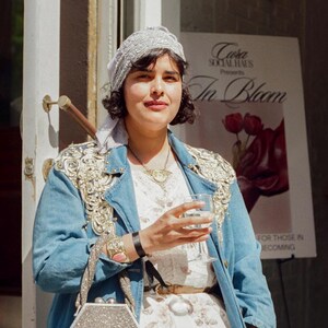 May include: A woman wearing a light blue denim jacket with gold embellishments, a white floral dress, and a silver hexagonal handbag. She is holding a glass of wine. A sign in the background reads "In Bloom."