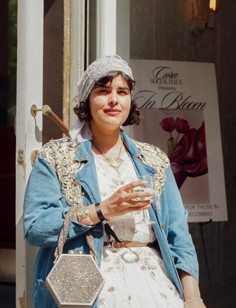 May include: A woman wearing a light blue denim jacket with gold embellishments, a white floral dress, and a silver hexagonal handbag. She is holding a glass of wine. A sign in the background reads "In Bloom."