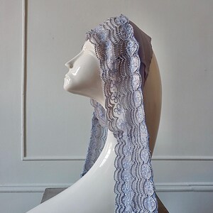 May include: A decorative head covering with intricate lace trim, displayed on a white mannequin head. The lace features a delicate floral pattern and is embellished with small, shiny beads. The head covering is a soft, muted blue color.