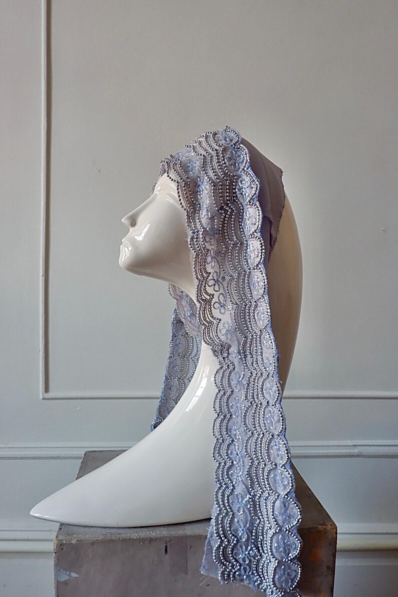 May include: A decorative head covering with intricate lace trim, displayed on a white mannequin head. The lace features a delicate floral pattern and is embellished with small, shiny beads. The head covering is a soft, muted blue color.
