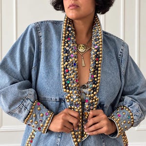 Puede incluir: Una chaqueta de mezclilla azul claro con solapas anchas y puños decorados con tachuelas metálicas doradas y pedrería redonda multicolor. La chaqueta tiene mangas largas y un cuello estructurado, mostrando un patrón denso de adornos metálicos.