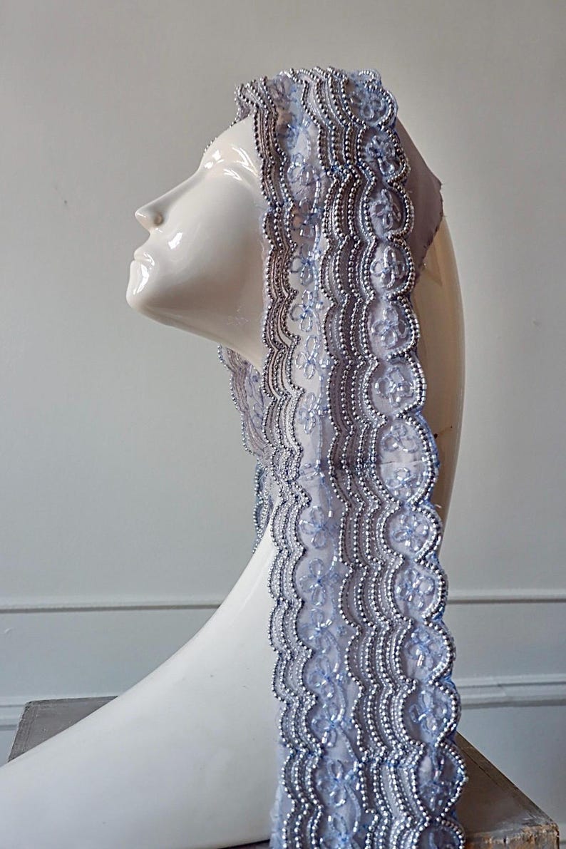May include: A light blue lace veil with silver beaded trim is draped over a white mannequin head. The veil's edge features a scalloped design. The background is a plain white wall.