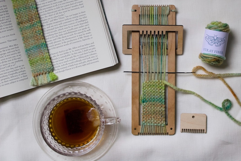 Może przedstawiać: Drewniane krosno tkackie z zieloną i ż&oacute;łtą przędzą w trakcie pracy, gotowa zakładka do książki i szpula przędzy "STRAY FIBER". Filiżanka herbaty i otwarta książka sugerują relaksującą aktywność rzemieślniczą.