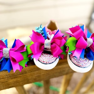 May include: Pink, blue, and green ribbon bows with rhinestone accents adorn a pair of white canvas sneakers. The toe caps are covered in pink rhinestones. The bows are attached to the laces of the shoes.