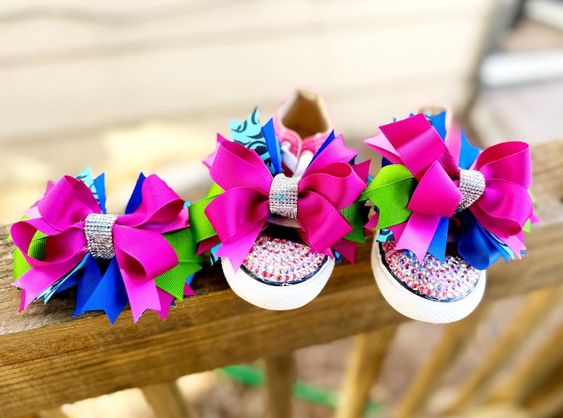May include: Pink, blue, and green ribbon bows with rhinestone accents adorn a pair of white canvas sneakers. The toe caps are covered in pink rhinestones. The bows are attached to the laces of the shoes.