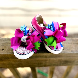 May include: A pair of pink canvas sneakers with large magenta bows and rhinestone embellishments on the toes. The bows feature blue, green, and patterned ribbons. The shoes are set against a wooden background.