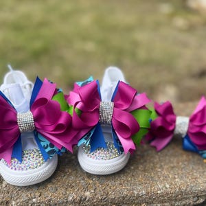 May include: White canvas sneakers adorned with large magenta, blue, and green bows, embellished with rhinestones. A matching bow is also present. The bows have a sparkling rhinestone center. The shoes are designed for children.