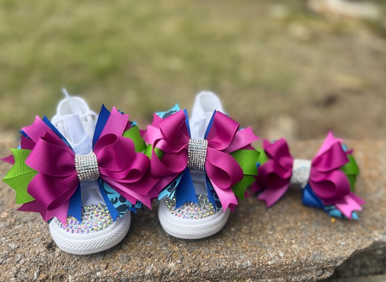 May include: White canvas sneakers adorned with large magenta, blue, and green bows, embellished with rhinestones. A matching bow is also present. The bows have a sparkling rhinestone center. The shoes are designed for children.