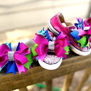 May include: Bright pink Converse sneakers with rhinestone accents and large, multi-colored bows. The bows feature fuchsia, blue, and green ribbons, with a sparkling rhinestone center. The shoes are set on a wooden surface.