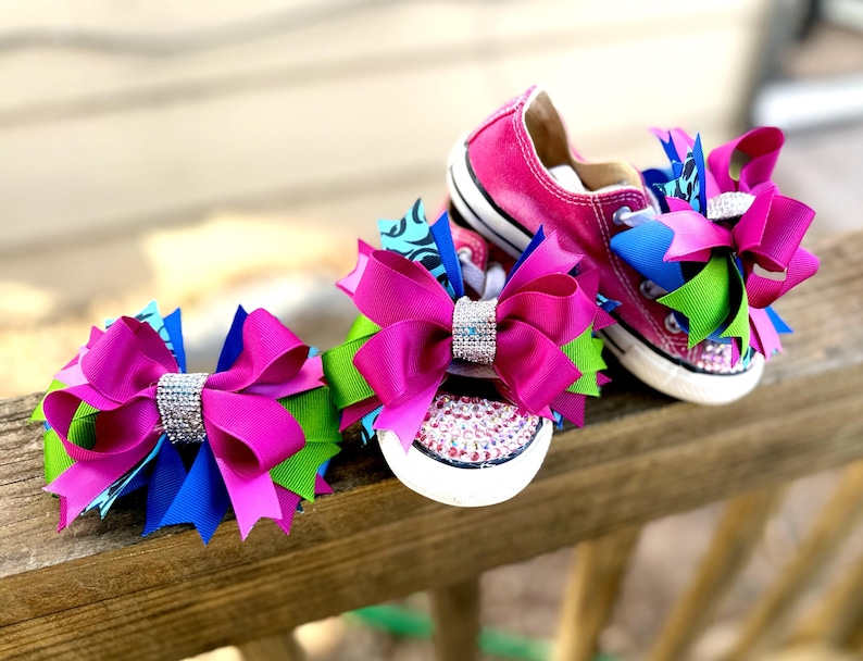 May include: Bright pink Converse sneakers with rhinestone accents and large, multi-colored bows. The bows feature fuchsia, blue, and green ribbons, with a sparkling rhinestone center. The shoes are set on a wooden surface.