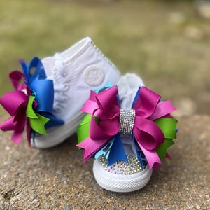 May include: A pair of white canvas sneakers with large decorative bows. The bows are a combination of fuchsia, blue, and green ribbons, with a rhinestone embellishment on one. The shoes are decorated with rhinestones.