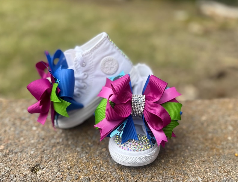 May include: A pair of white canvas sneakers with large decorative bows. The bows are a combination of fuchsia, blue, and green ribbons, with a rhinestone embellishment on one. The shoes are decorated with rhinestones.