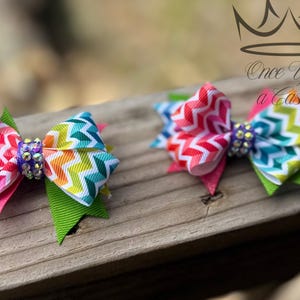 May include: Two colorful hair bows with a chevron pattern in rainbow colors. Each bow has a rhinestone center and is accented with pink and green ribbons. The bows are on a wooden surface.
