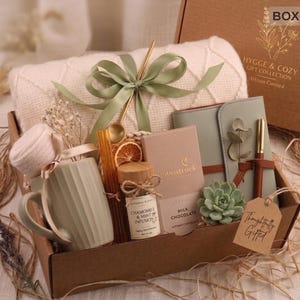 Op de afbeelding: Een geschenkdoos gevuld met een crèmekleurige deken, een saliegroene mok, een notitieboek, een pen en andere items. De doos bevat een label met de tekst "Thoughtfully Gifted". De doos is gelabeld "HYGGE & COZY GIFT COLLECTION".