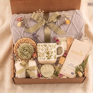 Puede incluir: Una caja de regalo llena de una manta gris, una taza con estampado floral, una barra de chocolate, una cuchara de madera, una vela y otros artículos de cuidado personal. La caja está atada con una cinta verde y decorada con flores secas. El texto "BOX 5" es visible.
