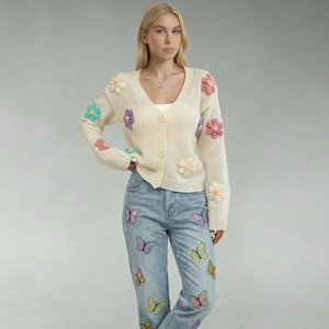 Puede incluir: Cárdigan color crema adornado con aplicaciones de flores 3D de colores. El suéter con cuello en V tiene cierre de botones. Los vaqueros azul claro con parches de mariposas completan el conjunto.