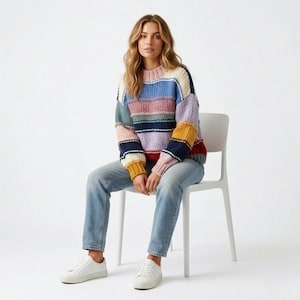Puede incluir: Un suéter a rayas multicolores con cuello redondo. El suéter presenta rayas horizontales en varios colores, incluyendo azul, rosa, amarillo y marino. La persona lleva vaqueros azul claro y zapatillas blancas. La persona está sentada en una silla blanca.
