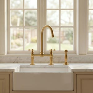 May include: A brushed gold kitchen faucet with a high-arch spout and two cross-style handles, accompanied by a matching side sprayer. The fixture is mounted on a white marble countertop behind a white farmhouse sink with a smooth finish.
