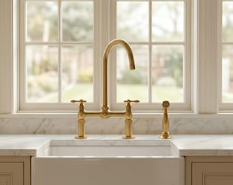 May include: A brushed gold kitchen faucet with a high-arch spout and two cross-style handles, accompanied by a matching side sprayer. The fixture is mounted on a white marble countertop behind a white farmhouse sink with a smooth finish.