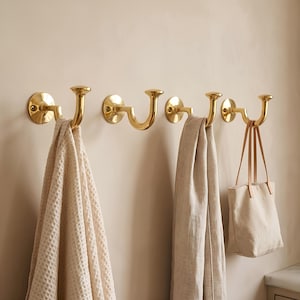 May include: Four polished brass wall hooks mounted in a horizontal row. Each hook features a circular base plate with visible screw holes and a curved arm ending in a rounded knob. A textured beige towel, a linen scarf, and a tote bag hang.