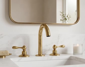 May include: A gold-toned bathroom faucet set with a curved spout and lever handles. The faucet is mounted on a white countertop with a marble pattern. A gold-framed mirror hangs above the sink, reflecting the faucet.