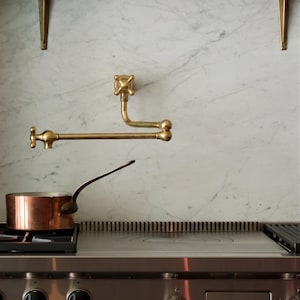 May include: A kitchen scene featuring a copper pot on a stainless steel stove. Above the stove is a brass faucet with a cross-shaped handle. The backdrop is a white marble wall with brass brackets.