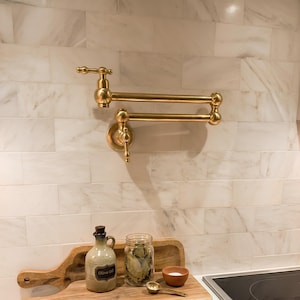 May include: A gold-colored, wall-mounted pot filler faucet with a two-jointed, articulated arm. The faucet is installed on a white marble tile backsplash. A wooden cutting board with a jar of bay leaves, a small bowl, and a bottle of olive oil are in the foreground.