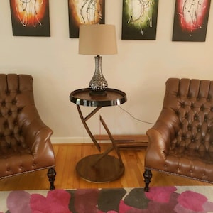 May include: Two brown leather tufted armchairs with dark wooden legs flank a round side table. The table features a dark circular top and a curved metal base. A table lamp with a textured silver base and a beige drum shade sits on the table.