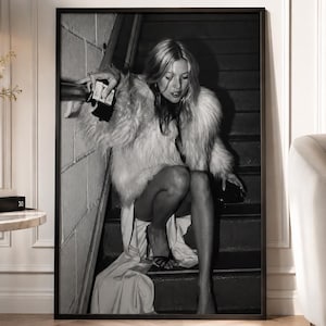 Könnte beinhalten: Ein schwarz-weiß gerahmter Wandkunstdruck, der eine Person mit langem Haar in einem strukturierten Pelzmantel und einem langen Rock zeigt, die auf einer Treppe sitzt und eine kleine Handtasche neben einem Treppengeländer vor einer Ziegelwand hält.