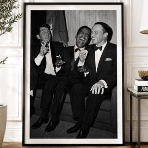 May include: A black and white framed photograph depicting three men in formal tuxedo suits sitting together on a sofa. The men are laughing and holding cigarettes while wearing bow ties and white dress shirts. The print is encased in a black frame.