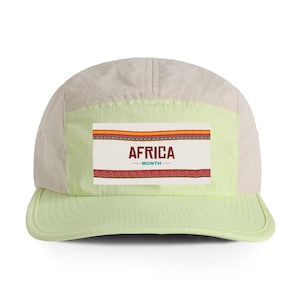 May include: A light green and beige five-panel cap with a rectangular patch. The patch features the text "AFRICA MONTH" in maroon, with decorative patterns in red, orange, and yellow.