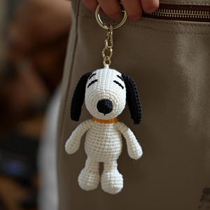 May include: A handmade crochet Snoopy keychain. The Snoopy figure is white with black ears and a black nose. It has a yellow collar and is attached to a gold-colored keychain.