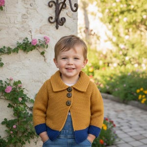 Puede incluir: Un cárdigan de punto amarillo mostaza con ribetes azul marino. El cárdigan tiene un cuello grande y tres botones marrones. El niño lleva un peto vaquero.