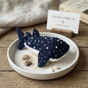 May include: A handmade crochet whale shark in navy blue with white accents and small white beads. The whale shark is displayed on a white plate with sand, small stones, and seashells. A business card reads "threads of light Art Handmade Crochet."