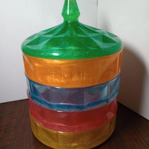 May include: A decorative jar with a faceted green lid and a stacked body of orange, blue, red, and gold. The jar has a glittery, iridescent finish, and a pointed finial on the lid. The jar is likely used for storage or display.